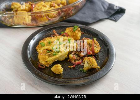 Cauliflower steaks baked with spices, herbs and red onions in a casserole, portion with parsley garnish on a black plate and a g Stock Photo