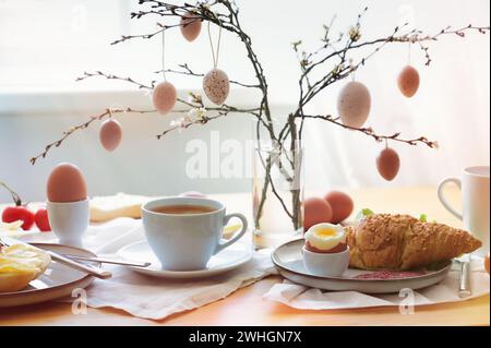 Easter breakfast in the morning sun with coffee and croissant on a table at the window, decoration from natural eggs hanging on Stock Photo