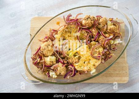 Baked cauliflower steaks with spicy oil and red onions in a glass casserole on a wooden kitchen board on a light table, vegetari Stock Photo