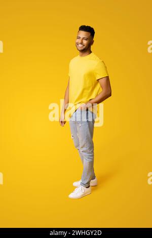 Pleased indian man in casual wear stands isolated on yellow, male student, young handsome guy standing in relaxed pose and looking aside, holding hand in pocket of jeans, full length Stock Photo