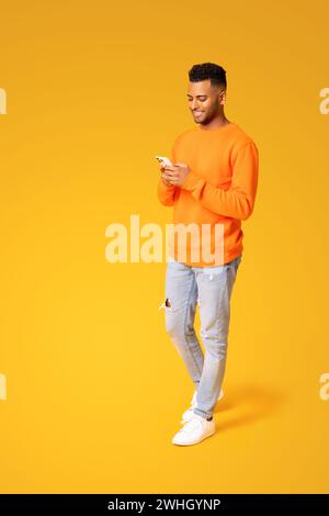 Full length photo of smiling young Indian man texting on the smartphone, using mobile phone for communication online, hindu male freelancer developer testing app on the phone isolated on yellow Stock Photo
