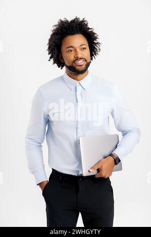 Smiling young African-American male entrepreneur stands in studio isolated on white and carrying trendy laptop. Cheerful black guy in formal wear holding closed laptop Stock Photo