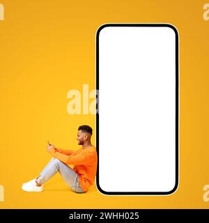 Arab man sitting near huge smartphone with empty blank screen, holding smartphone, enjoying chatting in social networks, texting messages, mockup, collage Stock Photo