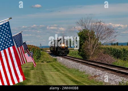 Restored Antique Steam Passenger Train Approaches a Fence With Gently Waving American Flags on It Stock Photo