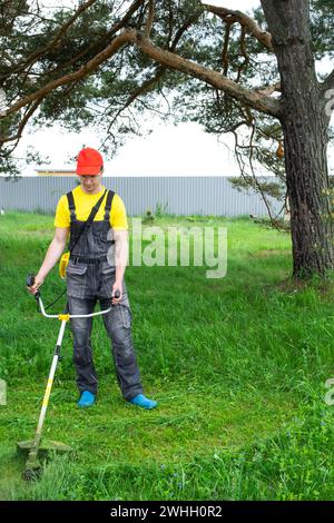 A male gardener mows the green grass of the lawn in the backyard with a gasoline mower. Trimmer for the care of a garden plot Stock Photo