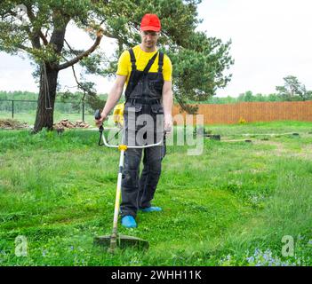 A male gardener mows the green grass of the lawn in the backyard with a gasoline mower. Trimmer for the care of a garden plot Stock Photo