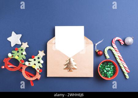 Christmas or Happy New Year envelope with white card and festive sparkling holiday decorations on blue background. Top view, copy space. Stock Photo