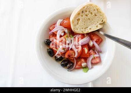 Fresh tomato salad with red onions, black olives and bread in a bowl on a white tablecloth, healthy snack, high angle view from Stock Photo