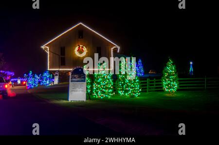 Festive Nighttime Scene Of A Building Outlined With White Christmas Lights And A Wreath, Surrounded By Trees Lit With Green Lights, Next To A Sign With Text And A Christmas Display. Stock Photo