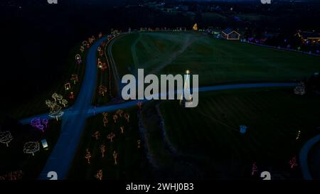 Aerial Nighttime View Of A Large Lit Christmas Tree And Multiple Smaller Trees Decorated With Colorful Lights Along A Winding Road. Stock Photo