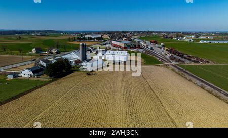 Aerial View Of A Farm Complex With Multiple Barns, Silos, And Plowed Fields In A Rural Setting Under A Clear Blue Sky. Stock Photo