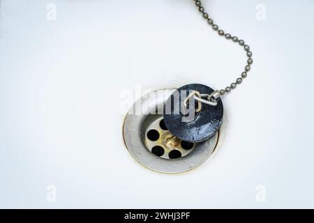 Old black rubber plug with metal chain on a rusty drain in a white wash basin or bathtub, copy space, selected focus Stock Photo