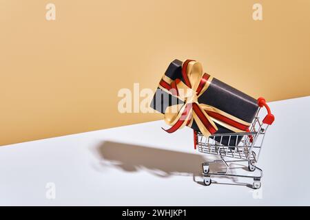 Shopping cart with gift box on beige gray background. Gifts wrapped in kraft black paper with ribbon and bow. Holiday Shopping c Stock Photo