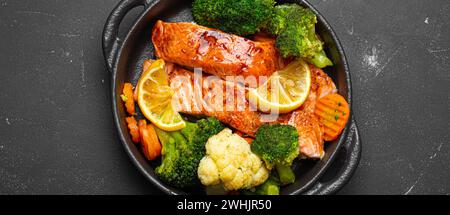 Healthy baked fish salmon steaks, broccoli, cauliflower, carrot in cast iron casserole bowl on black dark stone background. Cook Stock Photo