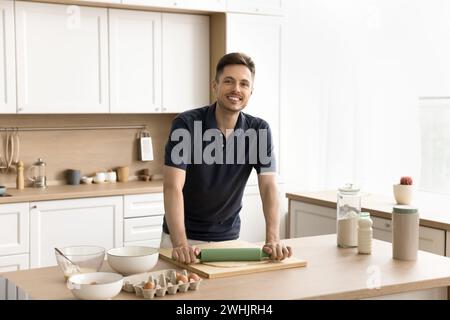 Cheerful handsome baker cooking at home, rolling dough Stock Photo