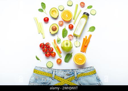 Fresh fruit, vegetables, smoothie falling into jeans and yellow measuring tape instead of belt on white background. Concept of h Stock Photo
