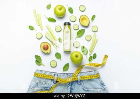 Green fruit, vegetables, smoothie falling into jeans and yellow measuring tape instead of belt on white background. Concept of w Stock Photo