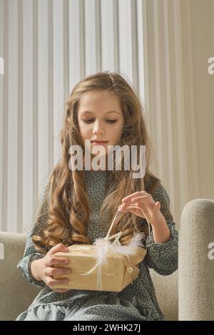 Portrait of young girl in a mint dress, sitting on a chair and pulling the ribbon of gift box Stock Photo