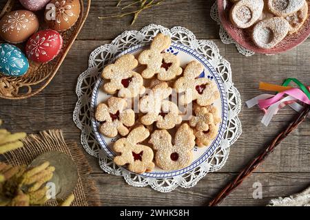 Homemade Linzer cookies in the shape of bunnies and chicken, with Easter eggs decorated with wax in the background. Top view. Stock Photo