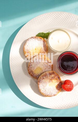 Four cheese pancakes with sour cream and strawberry jam on a white plate on a blue background. A delicious and healthy breakfast or dessert. Perfect f Stock Photo