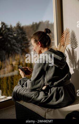 Woman in bathrobe sitting with mobile phone in a relaxation room during a wellness meal Stock Photo