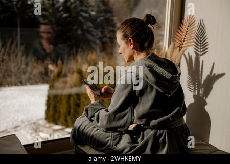 Woman in bathrobe sitting with mobile phone in a relaxation room during a wellness meal Stock Photo