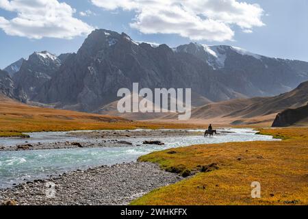 Rider riding through river in front of mountain landscape with yellow meadows, mountain peaks, Keltan Mountains, Sary Beles Mountains, Tien Shan Stock Photo