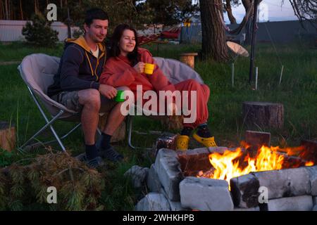 Couple in love is sitting by the outdoor campfire in the courtyard of the house on camping chairs, a romantic evening. a man and Stock Photo