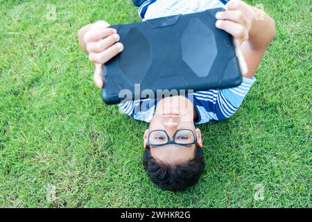 Teenager lies down on the lawn in the garden under the sun while using his tablet and wearing his glasses Stock Photo