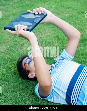 Teenager lies down on the lawn in the garden under the sun while using his tablet and wearing his glasses Stock Photo