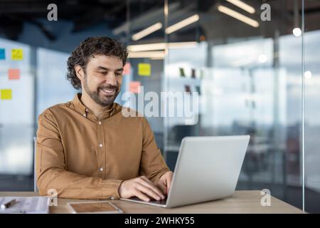 Mature experienced programmer inside office at workplace, businessman adult man typing on laptop keyboard, company worker in shirt satisfied with achievement results smiling. Stock Photo