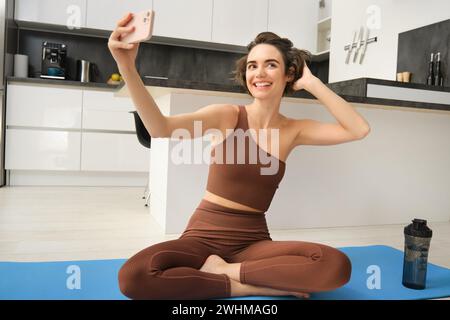 Lifestyle and workout. Modern fitness girl takes selfie on yoga mat, does her exercises at home and makes photos on smartphone, Stock Photo
