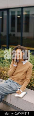 Cheerful business woman talking phone and making notes while sitting on building background Stock Photo