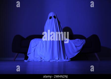 Creepy ghost. Woman covered with sheet sitting on sofa in blue light Stock Photo