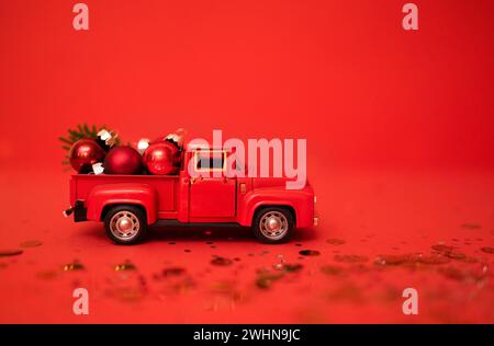 Red toy truck with Christmas balls and fir branch in its trunk. Miniature car with Christmas decoration on red background. Brigh Stock Photo