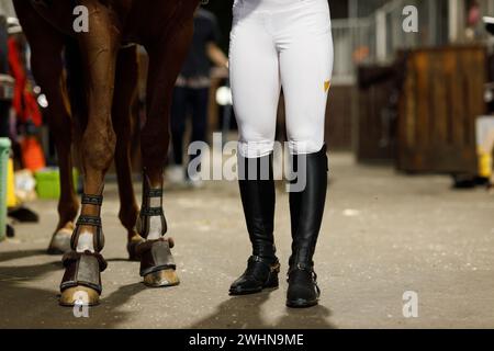 Female legs in black leather boots close up rider jockey walking with horse at stable and preparing horse racing or jumping comp Stock Photo