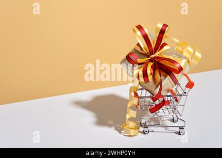 Shopping cart with gift box on beige gray background. Gifts wrapped in kraft paper with ribbon and bow. Holiday Shopping concept Stock Photo