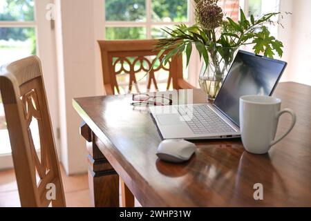 Laptop computer on a wooden dining table with coffee cup and flower bouquet in the living room, concept for home office working, Stock Photo
