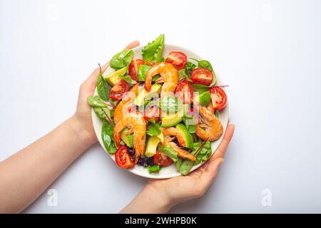 Female hands holding healthy salad with grilled shrimps, avocado, cherry tomatoes and green leaves on white plate isolated on wh Stock Photo