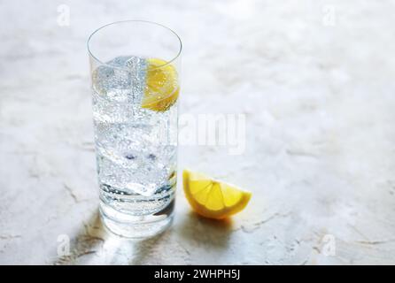 Fresh carbonated mineral water with ice cubes and lemon slices in a drinking glass, refreshing drink on a light stone background Stock Photo