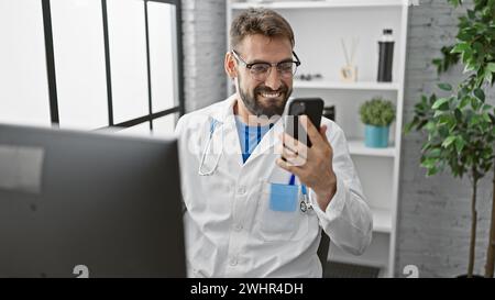 Smiling young hispanic man, a professional doctor, fervently types message on his smartphone while working at a medical clinic indoors Stock Photo