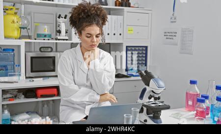 Hispanic woman working thoughtfully in a laboratory with microscope and laptop Stock Photo