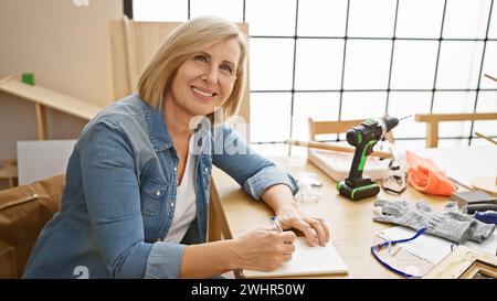 Mature blonde woman taking notes in a well-lit workshop with tools and woodwork Stock Photo