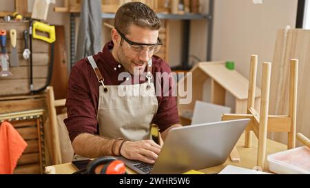 Handsome hispanic man with beard working on laptop in a carpentry workshop full of tools. Stock Photo