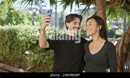 A smiling interracial couple takes a selfie together in a lush green park with tropical trees. Stock Photo