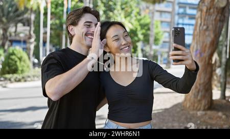 Happy interracial couple takes a selfie outdoors in a sunny city park, with green nature background. Stock Photo