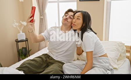An interracial couple takes a playful selfie in a well-lit bedroom, showing affection and togetherness in a cozy home setting Stock Photo