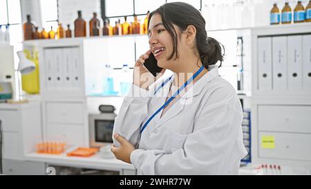 Young hispanic woman in lab coat speaking on phone in a modern laboratory indoor setting Stock Photo