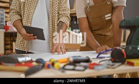 Two professional carpenters, a man and a woman, harness tech in their carpentry workshop, using a touchpad for drawing on paper amidst wood, tools, an Stock Photo