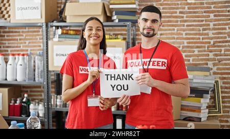 Confident young man and woman volunteers, standing in unity at charity center, holding a heartfelt 'thank you' paper banner in spotlight of altruism. Stock Photo